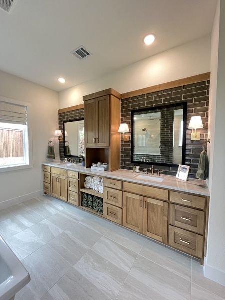 A modern bathroom with dual sinks, wood cabinetry, and elegant lighting, featuring a brick-style backsplash and tiled floor.
