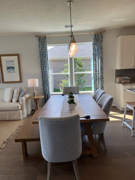 A cozy dining area with a wooden table, gray chairs, elegant pendant light, and large window with patterned curtains.