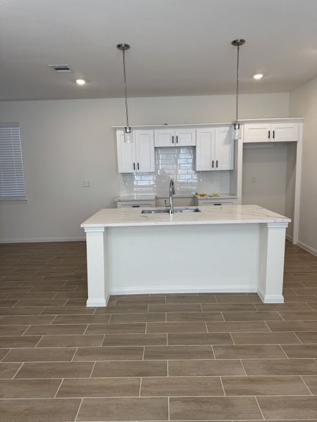 A modern kitchen with sleek white cabinetry, an island with a sink, pendant lights, and wood-look tile flooring.