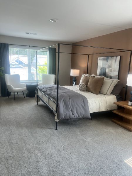 A cozy bedroom with a modern canopy bed, plush carpet, and natural light from large windows. Two accent chairs complete the space.