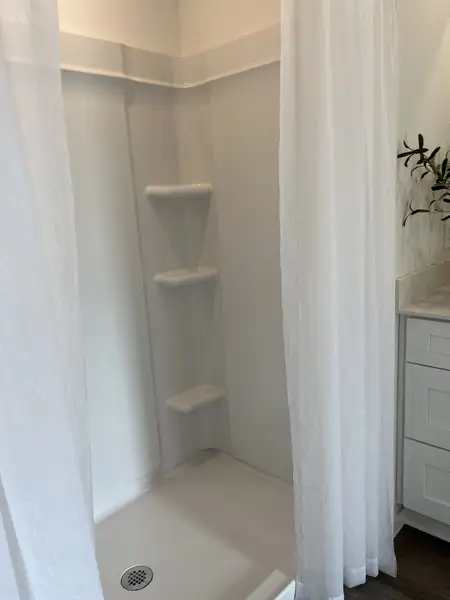 A sleek shower with corner shelving, white curtain, and adjacent vanity enhanced by greenery for a refreshing bathroom oasis.