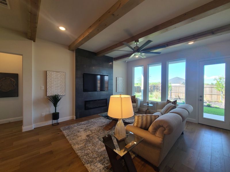 A cozy living room featuring a modern fireplace, exposed beams, and large windows with a view of the backyard.