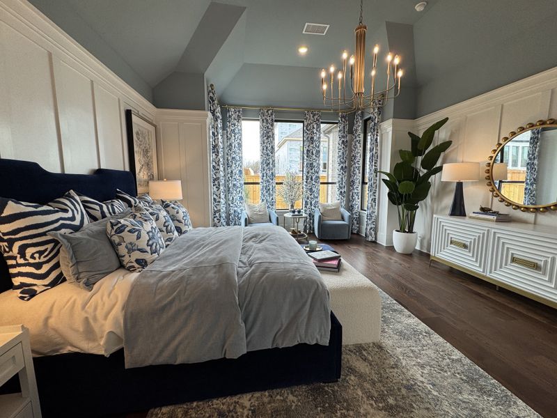 A luxurious bedroom with elegant decor, featuring a chandelier, patterned pillows, a plush bed, and stylish accent chairs.