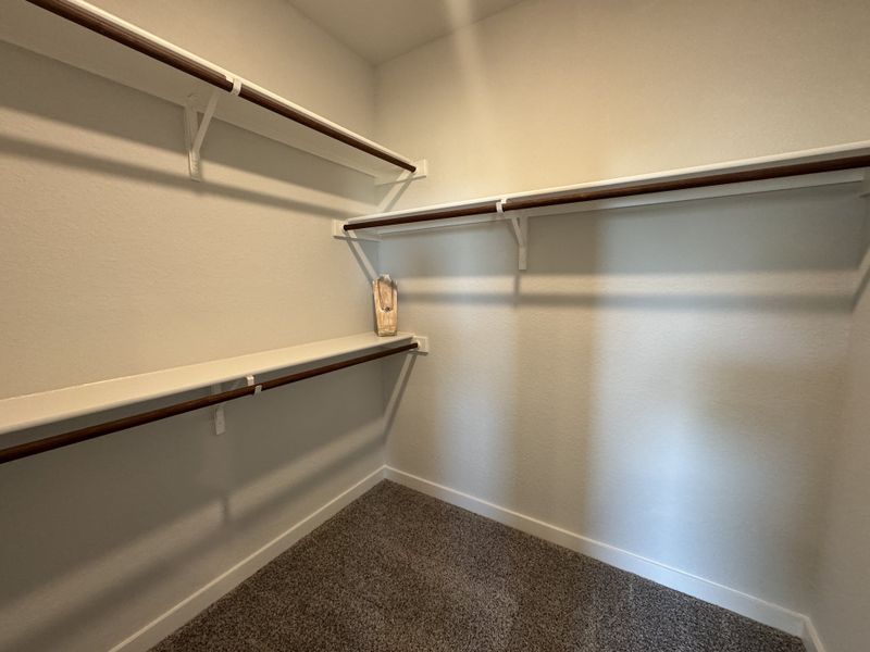 A spacious walk-in closet featuring white shelving, wooden rods, and plush carpeting. A spacious walk-in closet featuring white shelving, wooden rods, and plush carpeting.