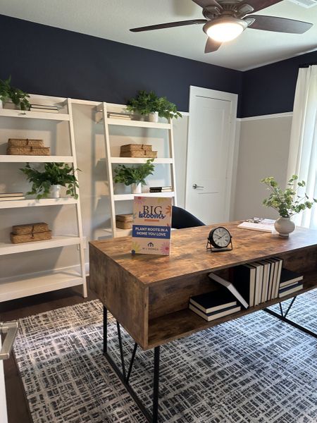A modern home office with a wooden desk, white shelving, and stylish decor featuring greenery, books, and a patterned rug. A modern home office with a wooden desk, white shelving, and stylish decor featuring greenery, books, and a patterned rug.