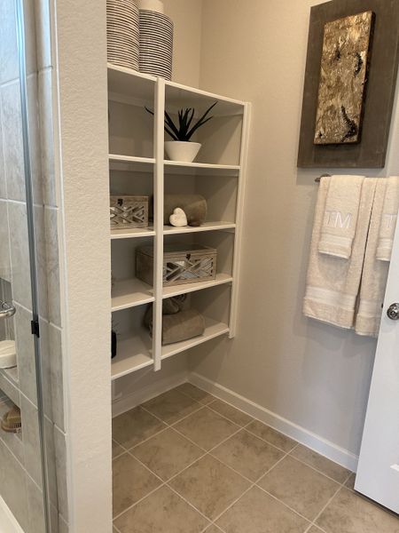 Elegant bathroom corner featuring open shelving, decorative items, and plush towels, with a neutral tile floor. Elegant bathroom corner featuring open shelving, decorative items, and plush towels, with a neutral tile floor.