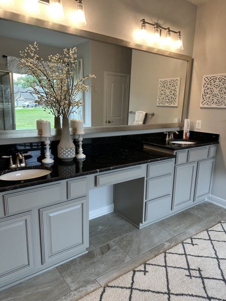 A bright bathroom featuring a long mirror, elegant lighting, gray cabinetry, and decorative accents. A bright bathroom featuring a long mirror, elegant lighting, gray cabinetry, and decorative accents.