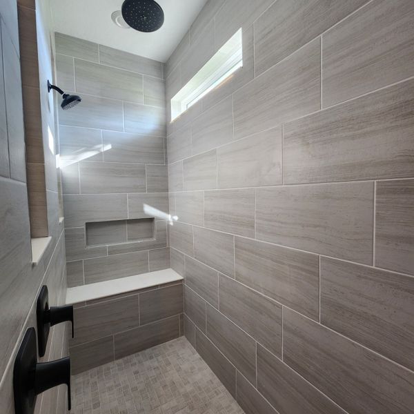 A sleek shower with large gray tiles, dual showerheads, and a built-in bench. Natural light filters through a small window.