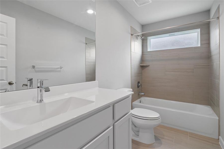 Secondary bath features tile flooring, bath/shower combo with tile surround, white stained wood cabinets, beautiful light countertops, mirror, dark, sleek fixtures and modern finishes.