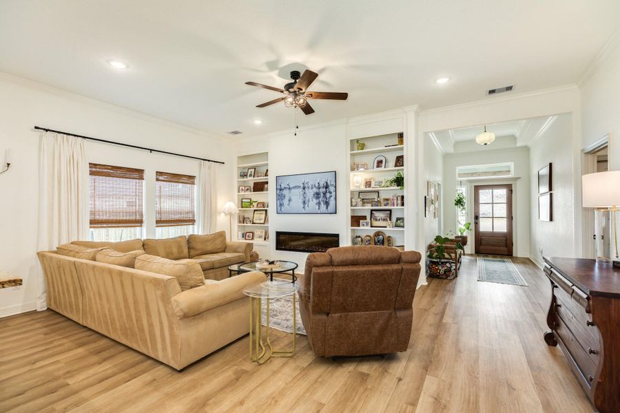 This inviting living room features an open layout, built-in shelving, and a wall-mounted TV. Natural light streams through large windows with wooden blinds, and the space is complemented by a ceiling fan and light wood look flooring.