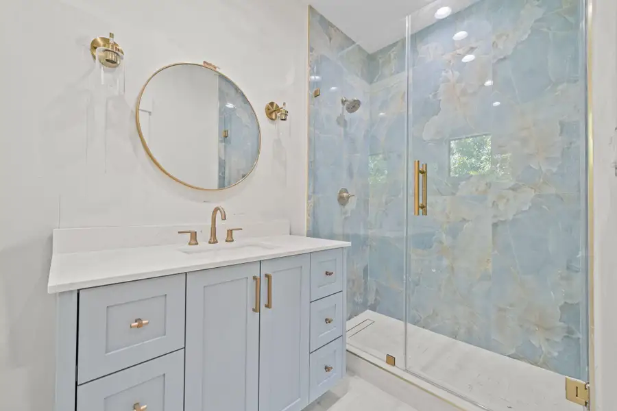 A sleek quartz countertop and sophisticated brushed-gold fixtures. The expansive frameless glass shower is enhanced by dramatic , marble-inspired wall tile with soft blue and white veining, creating a serene yet striking focal point. A refined round mirror and designer lighting complete the space, blending timeless elegance with contemporary luxury for a tranquil, resort style retreat.