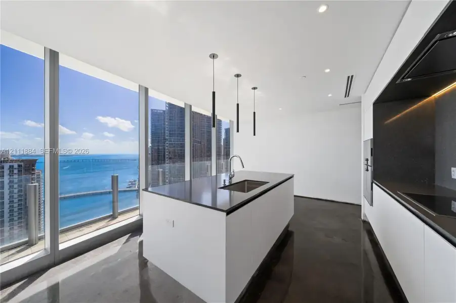 Kitchen featuring floor-to-ceiling windows, central island with an integrated stainless steel sink, polished concrete flooring, minimalist white cabinetry, and matte black pendant lighting Kitchen featuring floor-to-ceiling windows, central island with an integrated stainless steel sink, polished concrete flooring, minimalist white cabinetry, and matte black pendant lighting