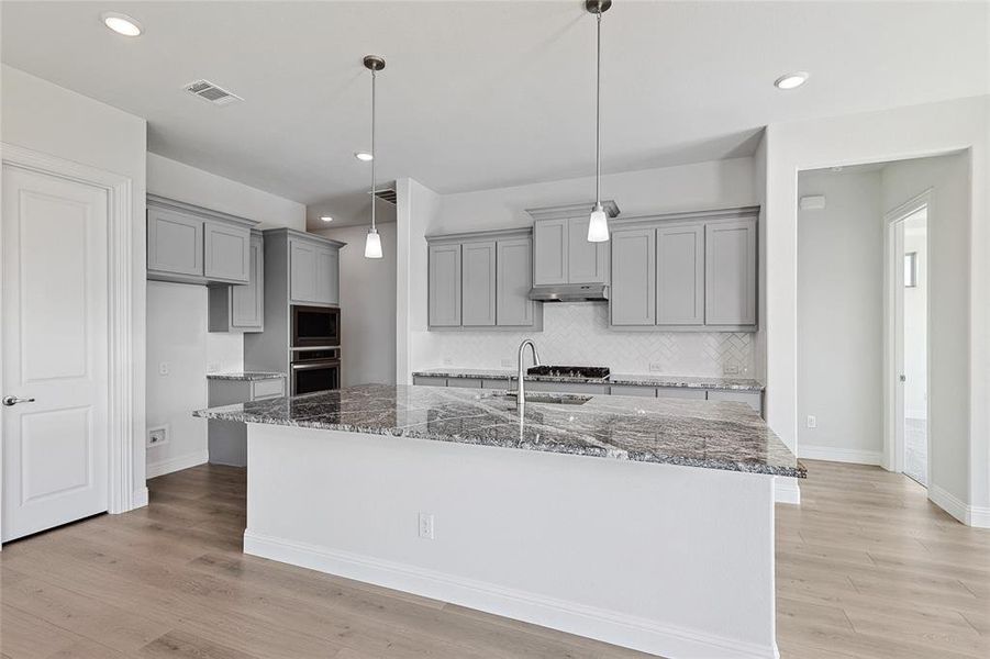 Kitchen featuring appliances with stainless steel finishes, a kitchen island with sink, dark stone counters, light wood-type flooring, and decorative light fixtures