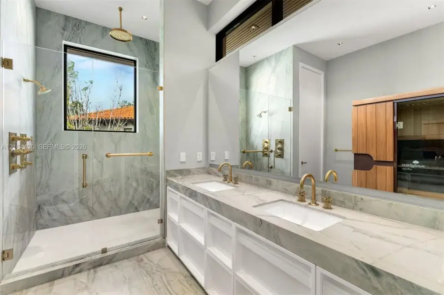 Bathroom featuring dual-sink vanity with stone countertop, walk-in shower with stone surround, brass-finish fixtures, and integrated sauna