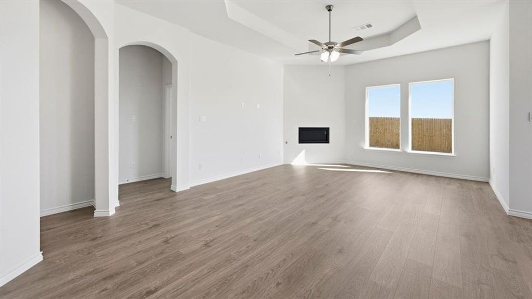 Spacious room featuring wood-finish flooring, a recessed electric fireplace, two large windows, a ceiling fan, and architectural arched doorways