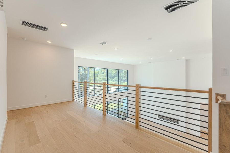 This photo shows a bright and airy loft space with light wood flooring and modern railing. Large windows provide ample natural light, and the space overlooks a lower level, creating an open and connected feel. Perfect for a home office or reading nook.