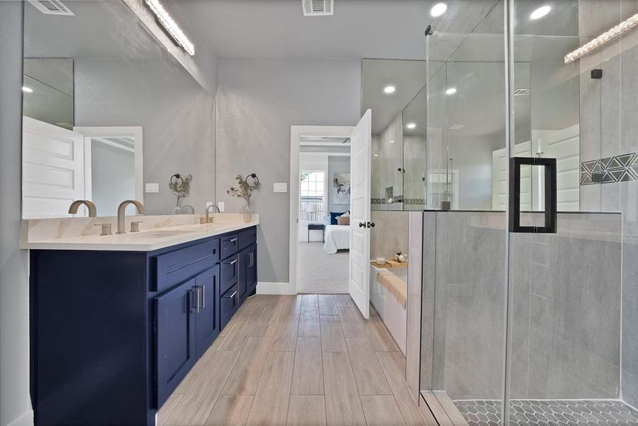 Bathroom featuring a double vanity with deep blue cabinetry and light-toned countertop, a frameless glass shower enclosure with hex tile flooring, and wood-finish flooring throughout