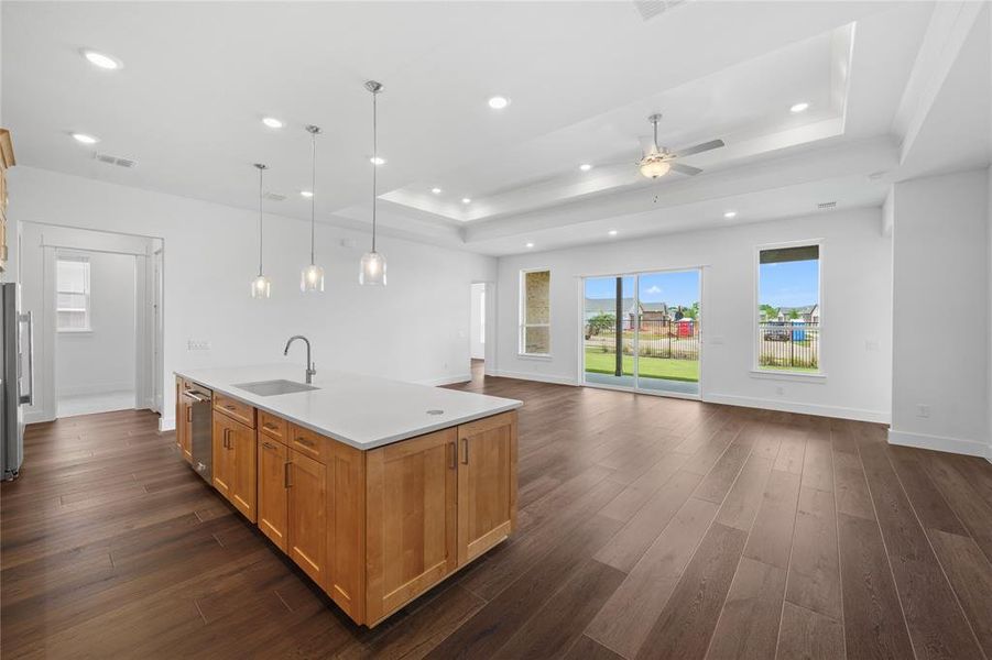 Spacious living area featuring dark wood-finish flooring, a tray ceiling with recessed lighting and a ceiling fan, and large windows with sliding glass doors opening to an outdoor patio