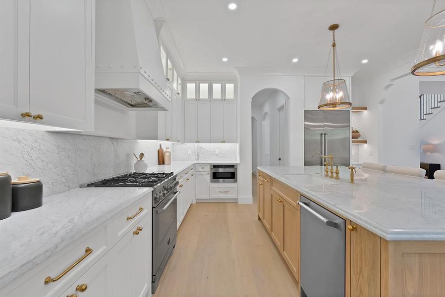 Gourmet kitchen featuring a substantial central island with a built-in sink and gold-tone faucet, white perimeter cabinetry with gold-tone hardware, and light wood-finish flooring