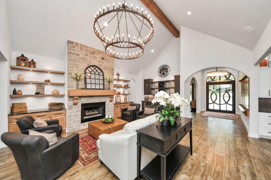 Elegant living space featuring a stunning stone fireplace, vaulted ceilings with exposed beams, and a grand chandelier. The open layout seamlessly connects to a welcoming entryway with a decorative door, creating a warm and inviting atmosphere.