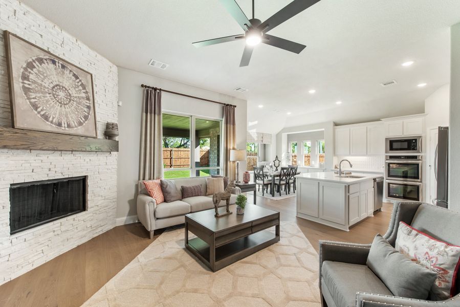 Open-concept living room with stone fireplace and ceiling fan, open to kitchen with white cabinets and island