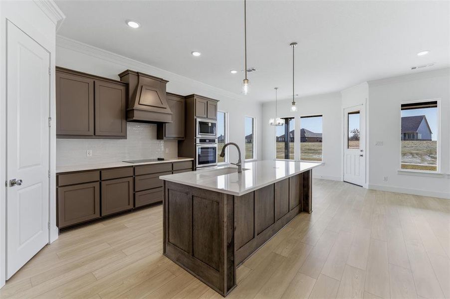 Kitchen with decorative backsplash, dark wood finish cabinetry, a center island with sink, light wood finished floors, and light stone counters