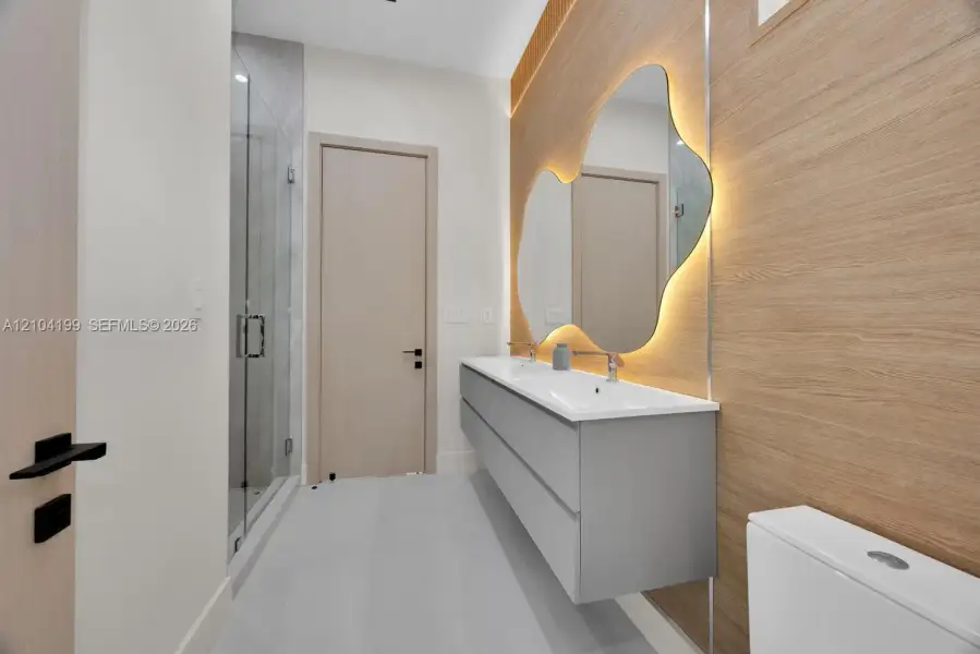 Floating grey vanity with integrated double sinks, oversized organic-shaped wall mirror with backlighting, wood-finish wall paneling, and glass-enclosed shower with tiled surround