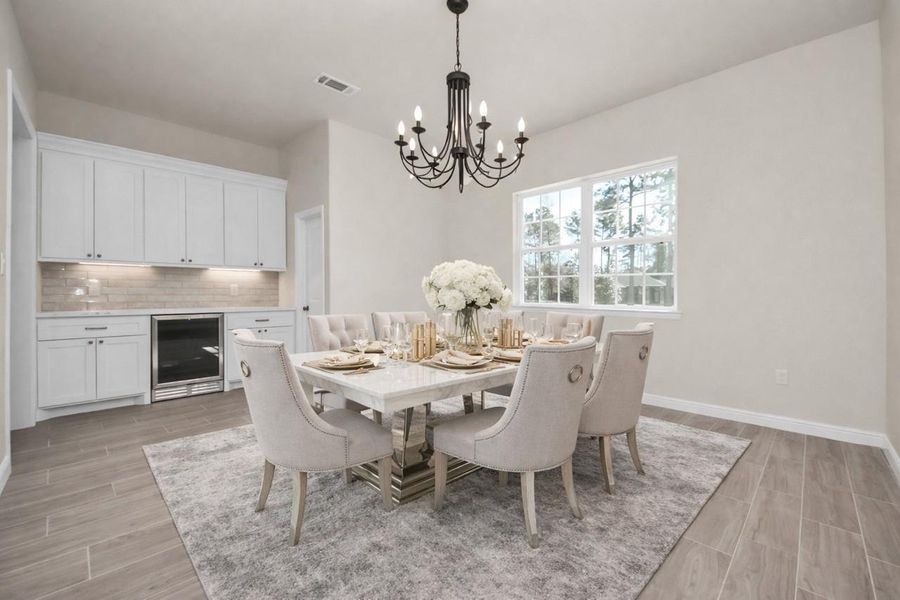 This virtually staged formal dining room features a modern chandelier and a large window providing natural light. The neutral tones and wood flooring create a warm, inviting atmosphere, complemented by built-in butler's pantry with custom cabinetry, a wine refrigerator, and a subtle backsplash.