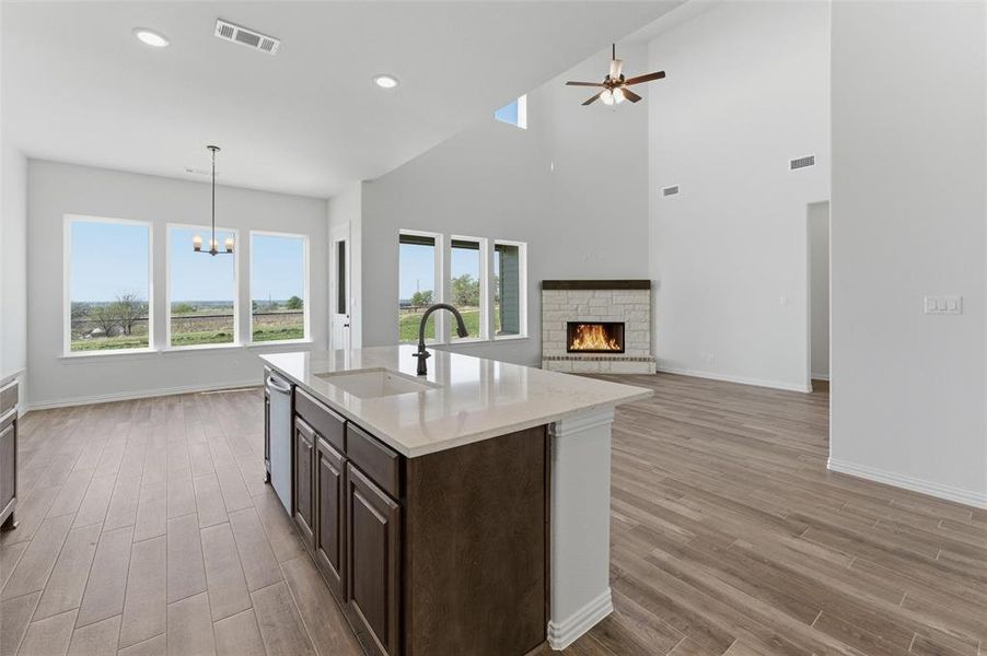 Kitchen with dark wood finish cabinetry, a warm lit fireplace, wood finish floors, a center island with sink, and light stone counters Kitchen with dark wood finish cabinetry, a warm lit fireplace, wood finish floors, a center island with sink, and light stone counters