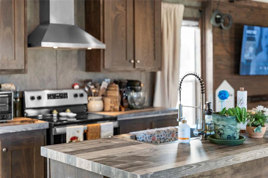Kitchen with wooden counters, wall chimney exhaust hood, dark brown cabinets, stainless steel electric range, and wood walls Kitchen with wooden counters, wall chimney exhaust hood, dark brown cabinets, stainless steel electric range, and wood walls