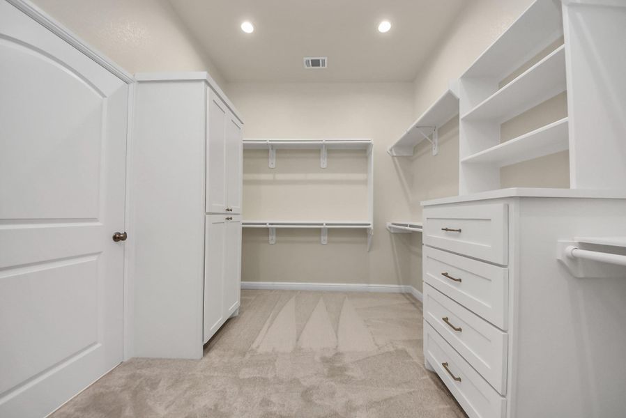 Spacious walk-in closet with ample shelving and hanging space, featuring white cabinetry and soft carpeted flooring. Bright and inviting, perfect for organized storage.