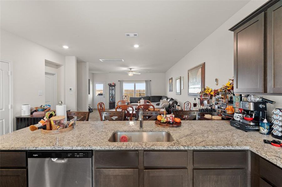 Kitchen with dark brown cabinetry, light stone countertops, stainless steel dishwasher, open floor plan, and a peninsula