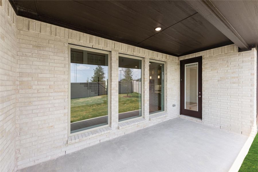 Covered back patio with light brick exterior, large windows, and dark-framed glass door leading inside