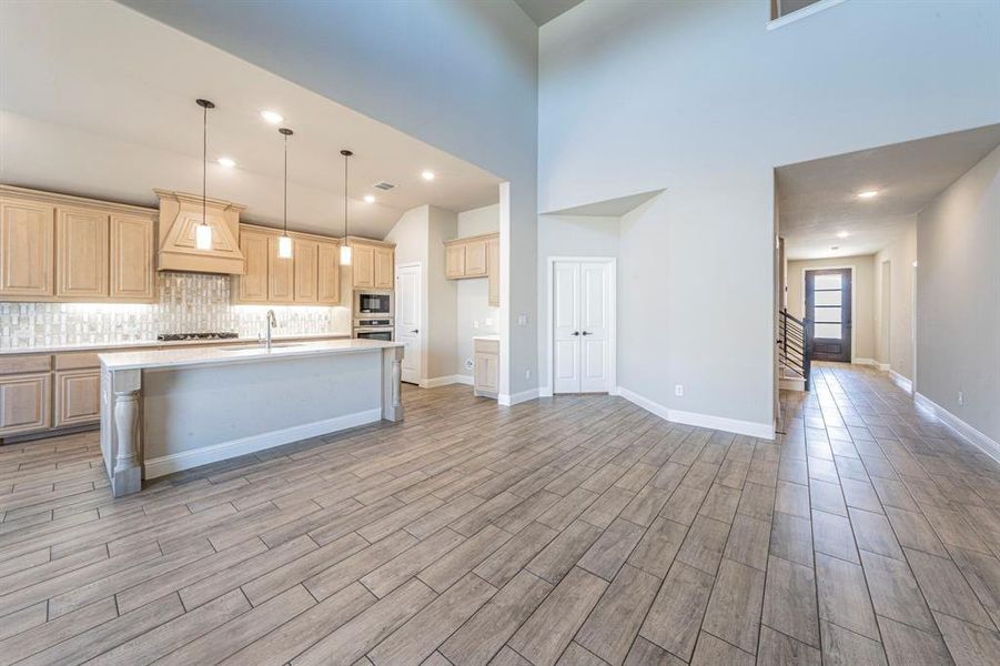 Spacious room featuring wood-finish flooring, a kitchen island with pendant lighting, light wood cabinetry, a tile backsplash, and high ceilings