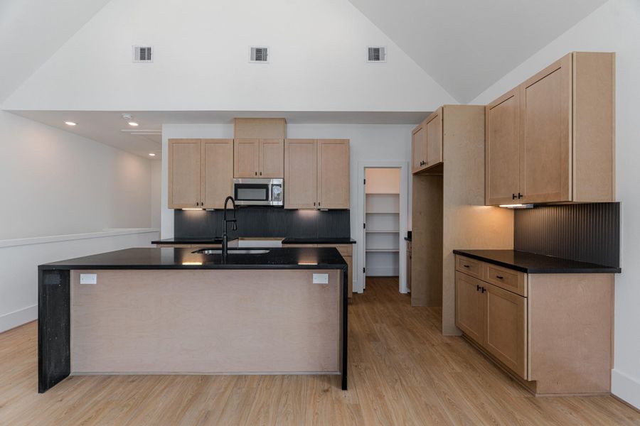 This kitchen features high ceilings, light wood cabinetry, and a spacious black countertop island with a sink. It includes modern appliances, ample storage, a walk-in pantry, and recessed lighting, all set against warm wood flooring.Photo Disclaimer: Photos are representative of the community and may depict a similar floor plan, model home, or previously completed residence. This kitchen features high ceilings, light wood cabinetry, and a spacious black countertop island with a sink. It includes modern appliances, ample storage, a walk-in pantry, and recessed lighting, all set against warm wood flooring.Photo Disclaimer: Photos are representative of the community and may depict a similar floor plan, model home, or previously completed residence.