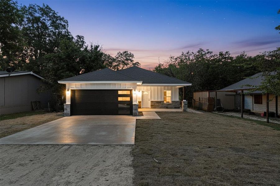 View of front facade with driveway, stone siding, a garage, a shingled roof, and a front yard