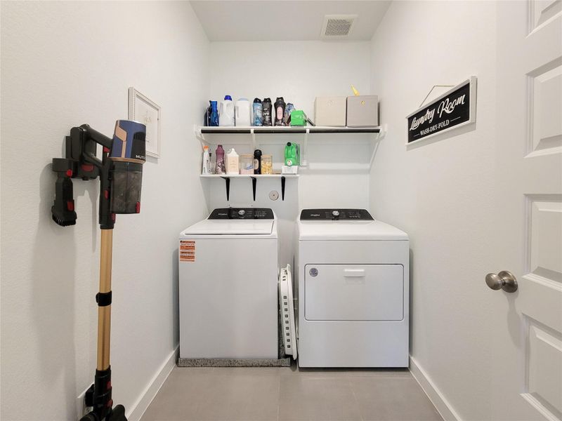This is a well-organized laundry room featuring a washer and dryer with shelving above for storage. The room is compact and functional, with cleaning tools neatly arranged on one side.