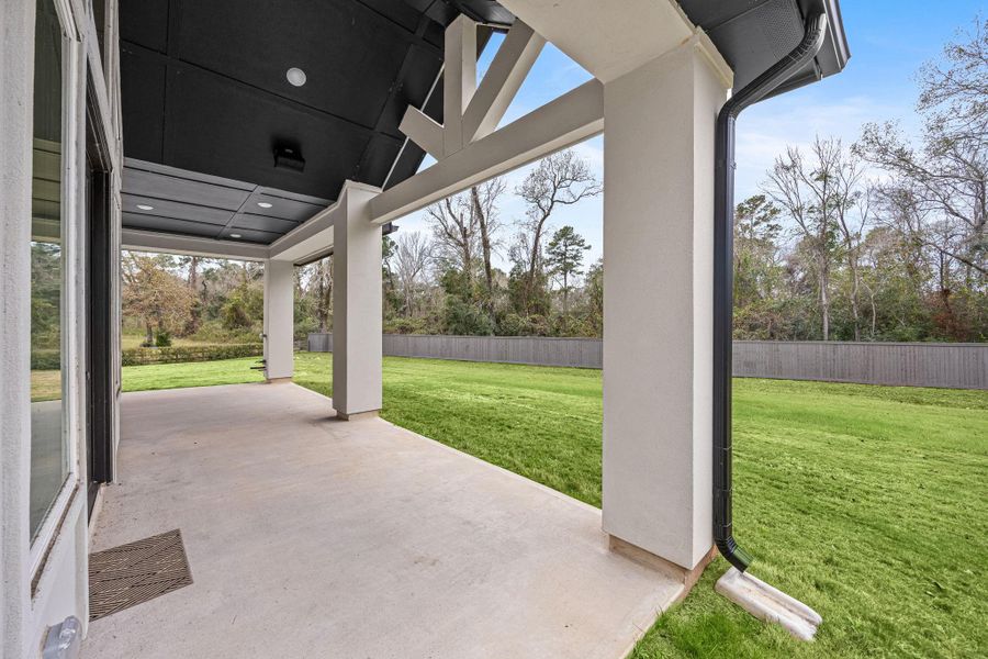 This photo showcases a spacious covered patio with a modern design, overlooking a large, grassy backyard enclosed by a wooden fence, offering privacy and a serene view of trees.
