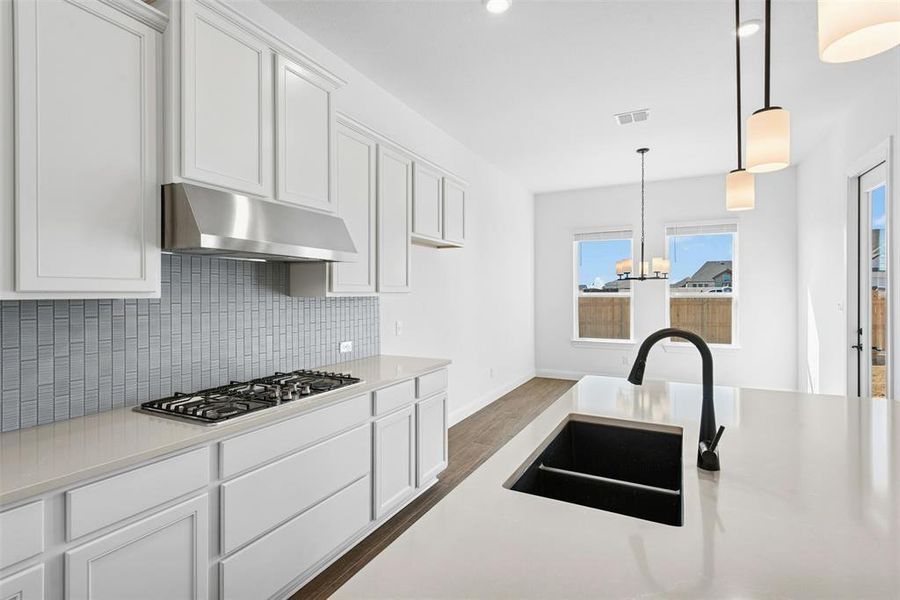 Modern kitchen featuring white shaker cabinetry, light-toned countertops, a gas cooktop with stainless steel hood, and a dark basin sink with a matte black faucet