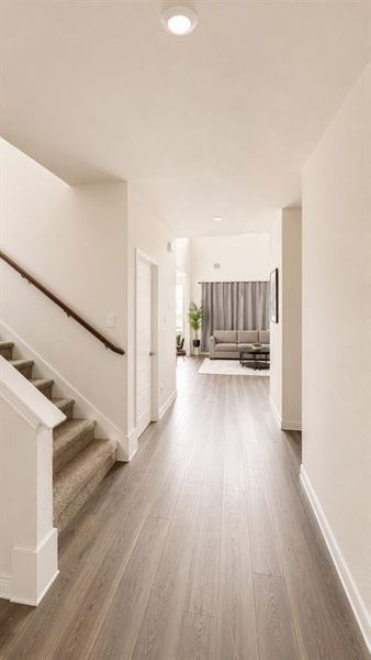 Hallway featuring wood-finish flooring, recessed lighting, white baseboards, and a carpeted staircase with a wood handrail Hallway featuring wood-finish flooring, recessed lighting, white baseboards, and a carpeted staircase with a wood handrail