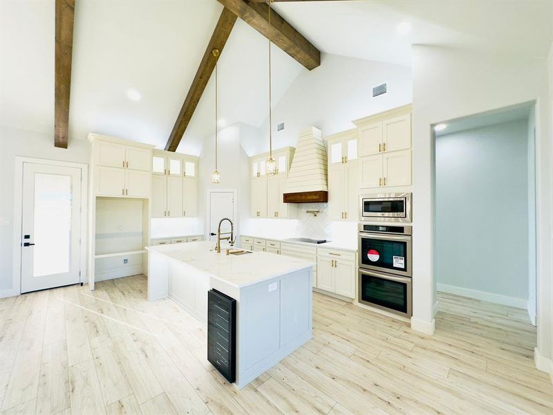 Kitchen with stainless steel appliances, wine cooler, premium range hood, decorative light fixtures, and a kitchen island with sink