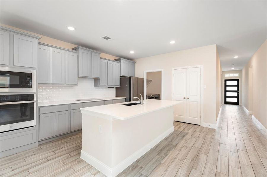Kitchen with gray cabinets, stainless steel appliances, light wood-style flooring, a center island with sink, and tasteful backsplash