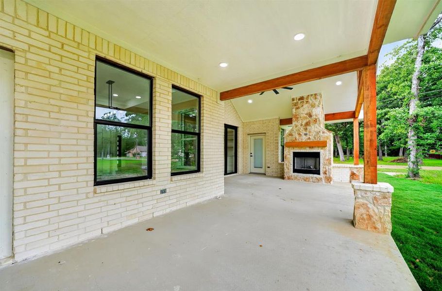 Expansive covered patio featuring a stone fireplace with a wood-finish mantle, exposed wood beams, brick exterior, and recessed lighting Expansive covered patio featuring a stone fireplace with a wood-finish mantle, exposed wood beams, brick exterior, and recessed lighting