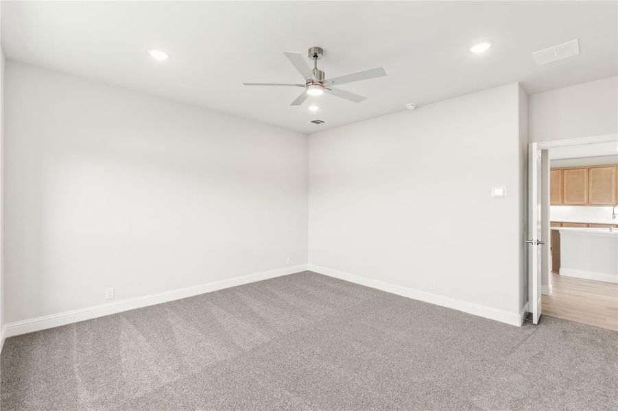 Empty bedroom with gray carpet, ceiling fan, white walls, and open door to bathroom with wood cabinets