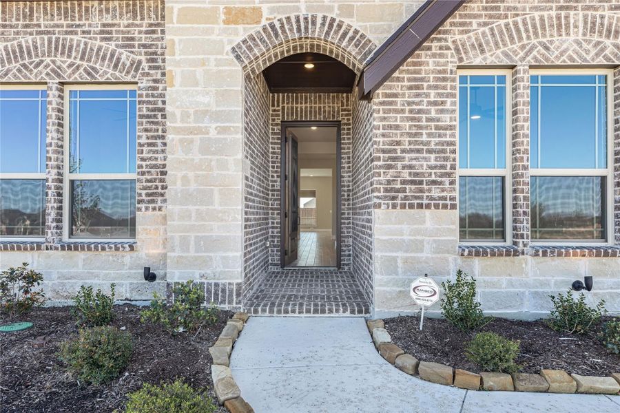 Front entry of a new home with arched brick doorway, stone exterior, and concrete walkway with landscaped beds Front entry of a new home with arched brick doorway, stone exterior, and concrete walkway with landscaped beds