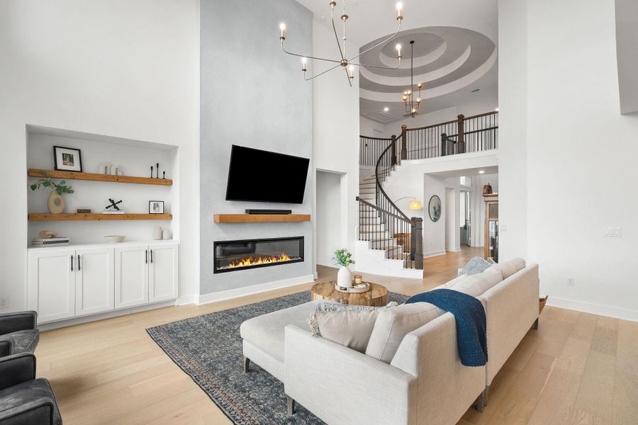 Grand living space featuring a two-story ceiling, light wood-finish flooring, and a linear electric fireplace with a floating wood mantel Grand living space featuring a two-story ceiling, light wood-finish flooring, and a linear electric fireplace with a floating wood mantel