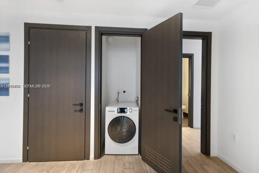 Laundry closet featuring a built-in front-load washer, dark wood-finish interior doors with matte black hardware, and wood-finish flooring Laundry closet featuring a built-in front-load washer, dark wood-finish interior doors with matte black hardware, and wood-finish flooring