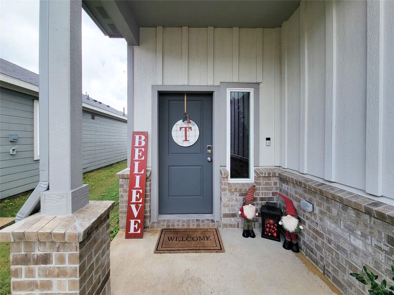 Charming front porch with a welcoming atmosphere, featuring a dark gray door, decorative signage, and festive gnomes. The brick and wood paneling add a cozy touch.