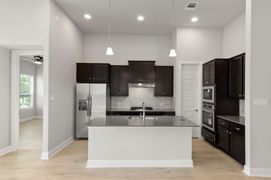 Kitchen with stainless steel appliances, backsplash, a center island with sink, a ceiling fan, and light wood-type flooring