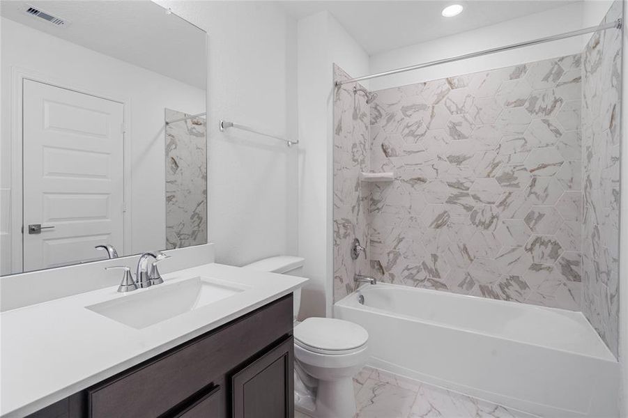 Secondary bath features tile flooring, bath/shower combo with tile surround, stained wood cabinets, beautiful light countertops, mirror, dark, sleek fixtures and modern finishes.